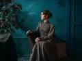 Elegancka kobieta w sukni w stylu light academia siedzi w fotelu, wpatrzona w dal.
