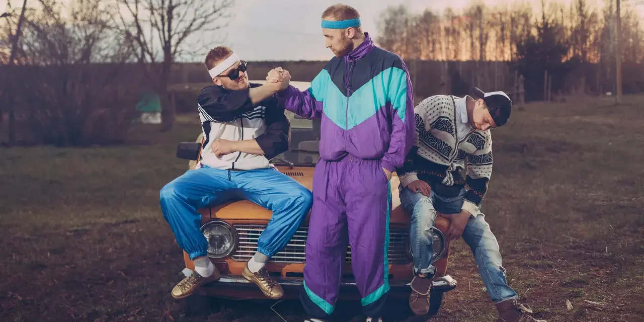 Trzech mężczyzn w dresach i swetrach, charakterystyczna moda lat 90. Siedzą na masce starego samochodu.