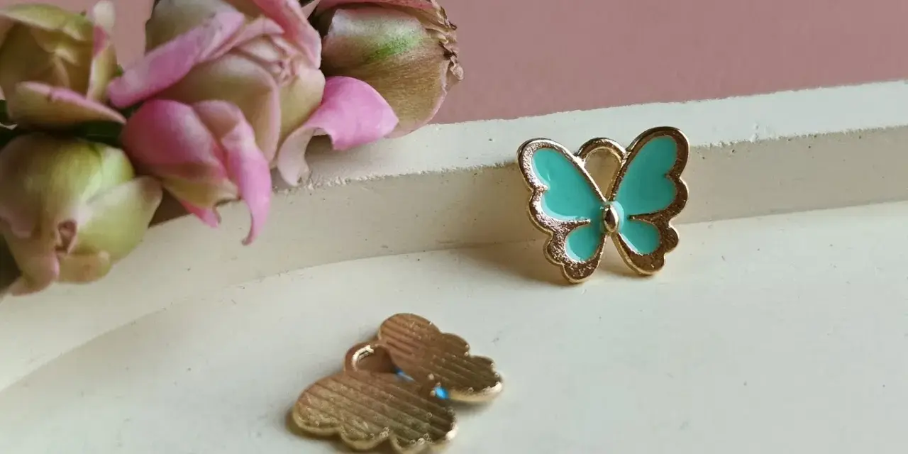 Dwa złote charms motyl, jeden turkusowy, drugi z widocznym wzorem. W tle delikatne pąki kwiatów.