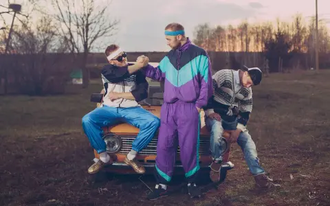 Trzech mężczyzn w dresach i swetrach, charakterystyczna moda lat 90. Siedzą na masce starego samochodu.