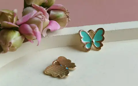 Dwa złote charms motyl, jeden turkusowy, drugi z widocznym wzorem. W tle delikatne pąki kwiatów.