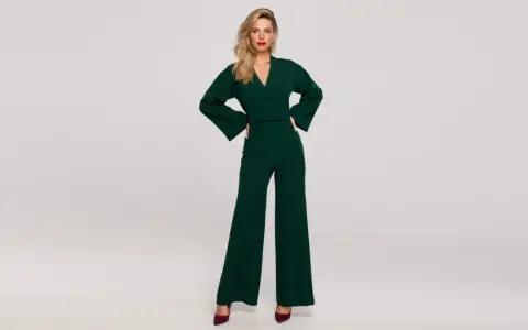 Elegancka kobieta w zielonym kombinezonie z szerokimi nogawkami. Góra kombinezonu ma dekolt kopertowy i szerokie rękawy.