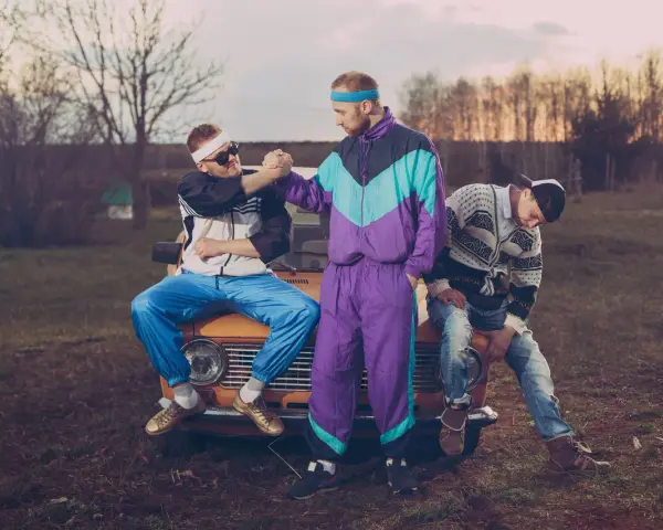 Trzech mężczyzn w dresach i swetrach, charakterystyczna moda lat 90. Siedzą na masce starego samochodu.