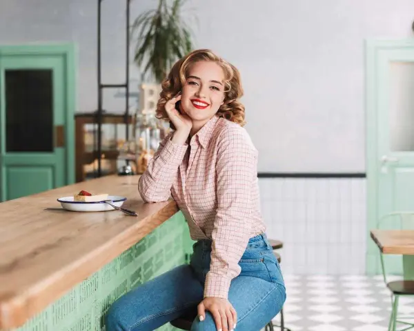 Uśmiechnięta blondynka w koszuli w kratę i dżinsach, stylizacja inspirowana modą PRL lat 70.