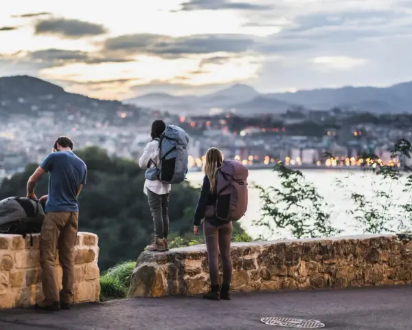 Trzy osoby z plecakami, gotowe na miejską wycieczkę, podziwiają panoramę miasta o zachodzie słońca.