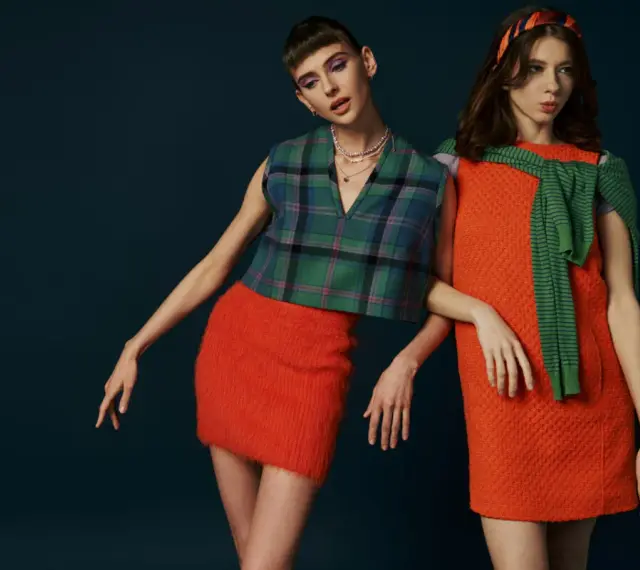 Dwie modelki w stylizacjach inspirowanych modą lat 60. Jedna w kraciastej kamizelce i pomarańczowej spódniczce, druga w pomarańczowej sukience i zielonym sweterku.