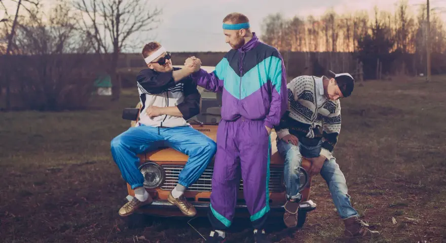 Trzech mężczyzn w dresach i swetrach, charakterystyczna moda lat 90. Siedzą na masce starego samochodu.