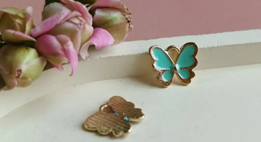 Dwa złote charms motyl, jeden turkusowy, drugi z widocznym wzorem. W tle delikatne pąki kwiatów.