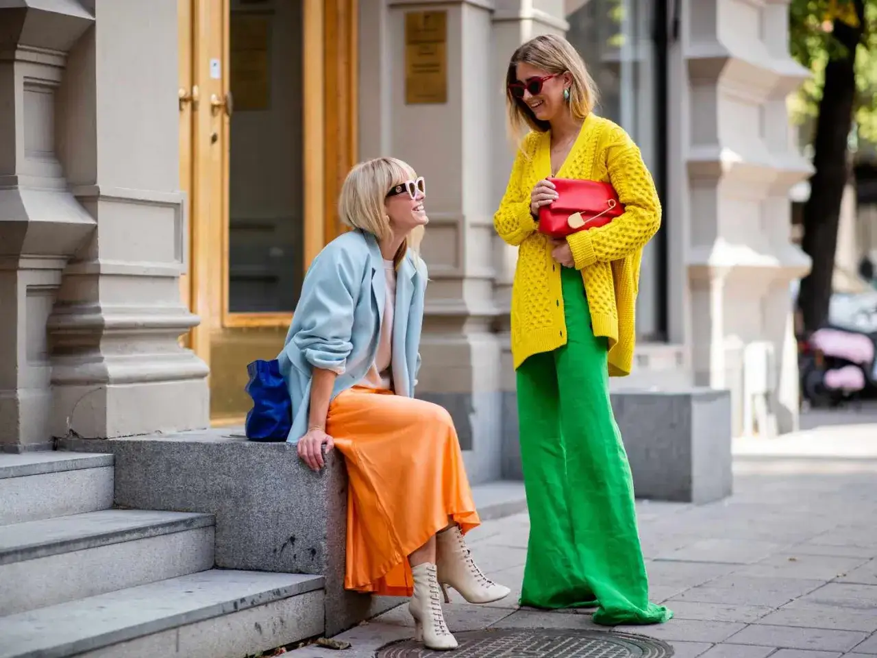 Dwie kobiety prezentują odważny styl color blocking: jedna w błękitnej marynarce i pomarańczowej spódnicy, druga w żółtym swetrze i zielonych spodniach.