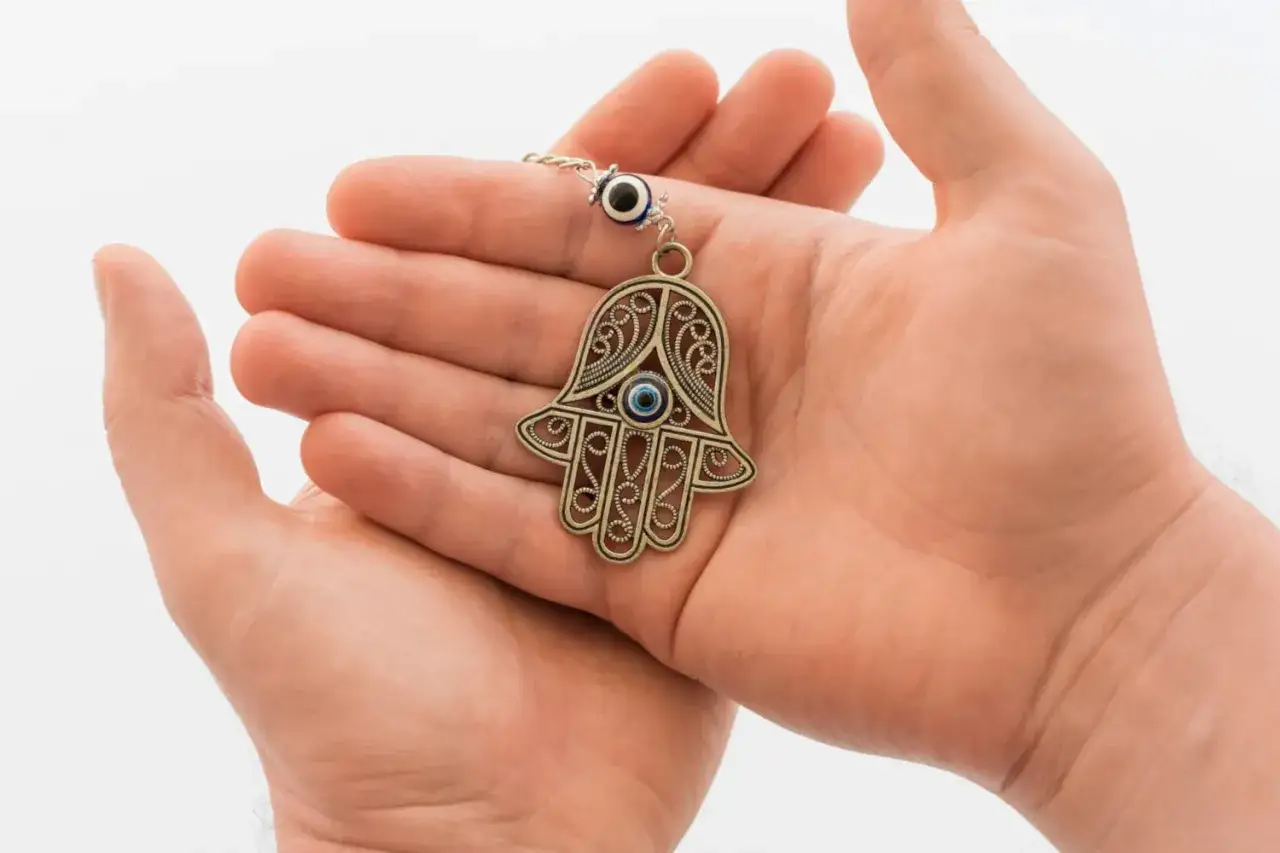 Dłonie trzymają ozdobny amulet w kształcie dłoni z okiem proroka, symbolizujący rękę opatrzności.