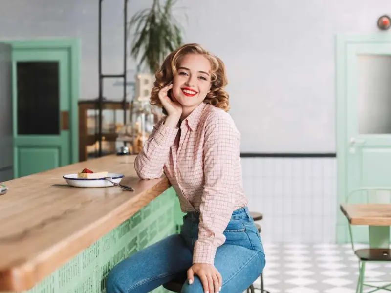Uśmiechnięta blondynka w koszuli w kratę i dżinsach, stylizacja inspirowana modą PRL lat 70.